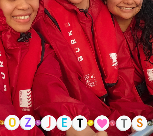 Three young women posing for a selfie on the jet boat in red ponchos and life jackets.